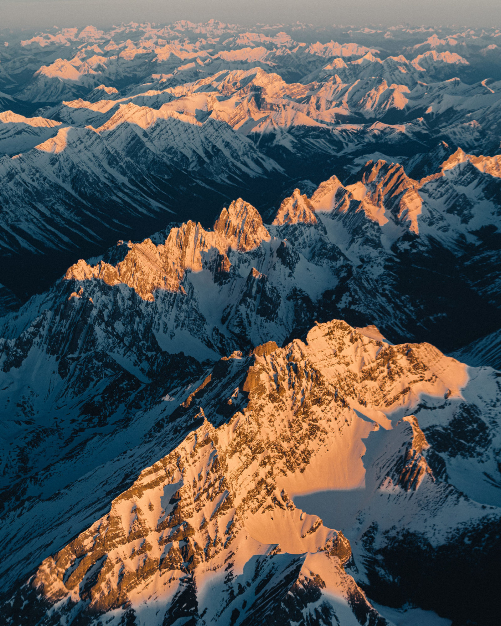 Canadian rocky mountains in Banff covered in snow lit up in a warmth of golden glowing light from the morning sunrise sun. Seen from an aerial perspective looking down at the mountains below. Photo taken by Michael Frymus