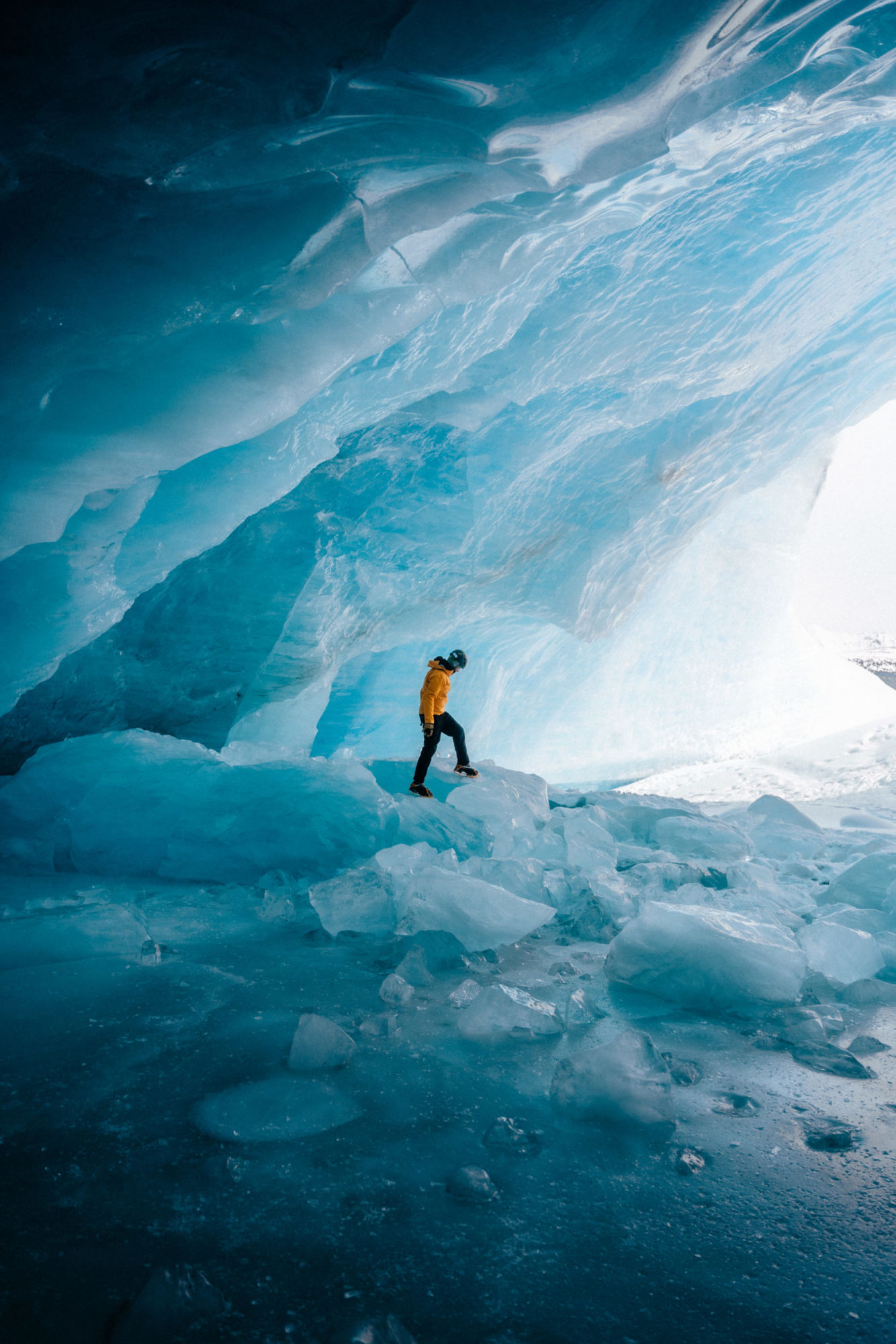 A man in a warm yellow jacket standing inside a cold blue, glass-like ice cave. Photo taken by Michael Frymus